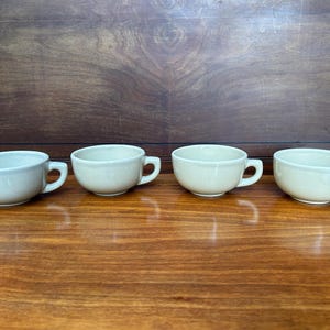 May include: Four light beige ceramic coffee mugs with rounded handles. The mugs are arranged in a row on a wooden surface.