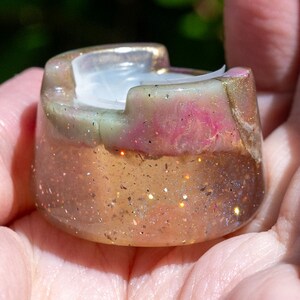 May include: A small, circular resin holder featuring a translucent base filled with gold glitter and a top section marbled with swirls of pink, green, and white. The object has a notched rim and a smooth, polished finish throughout its structure.