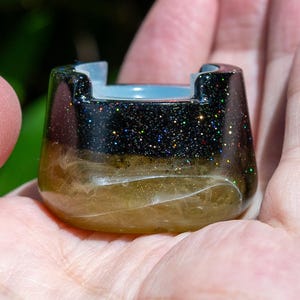 May include: A small, bowl-shaped object with a smooth, glossy finish. The top section is black with fine, multicolored glitter particles, while the bottom section features a translucent, amber-toned resin with swirling, cloud-like patterns.