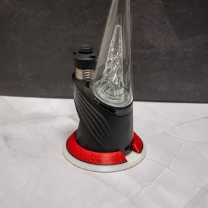 May include: A black electronic vaporizer device featuring a clear glass conical top with an internal spiral percolator. The base is matte black with curved ridges, accented by a metallic red ring and a white circular stand at the bottom.