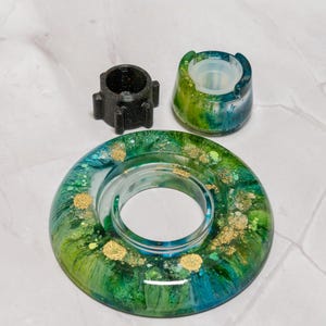 May include: A circular resin coaster with a swirling green, blue, and gold glitter pattern, accompanied by a small black plastic component with four protruding tabs and a matching green and blue resin holder with a translucent silicone insert.