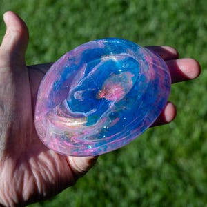 May include: A round, shallow resin dish featuring a swirling marbled pattern of translucent blue and pink hues. The surface is embedded with fine glitter particles and small gold-colored flakes, creating a glossy, reflective finish with depth.