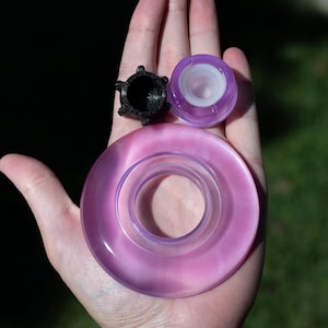 May include: A set of three components including a large, translucent, circular, light purple ring, a smaller, translucent, light purple circular piece with a white inner rim, and a small, black, textured, star-shaped cap with five points.
