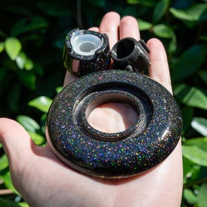 May include: A black circular object with a holographic glitter finish, featuring a central opening and a smooth, reflective surface. Two smaller, matching black glitter components with translucent white inserts are positioned above the main ring.