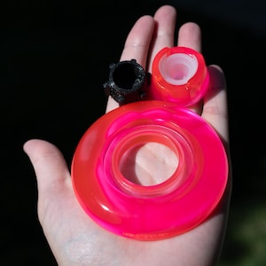 May include: A set of three small components including a large, flat, circular neon pink ring, a smaller neon pink cylindrical piece with a translucent white center, and a textured black plastic part with a central opening and small protrusions.