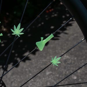 May include: Light green, glow-in-the-dark plastic spoke beads shaped like cannabis leaves and a water pipe attached to the thin black metal spokes of a bicycle wheel. The decorative accessories are arranged individually along the wheel spokes.