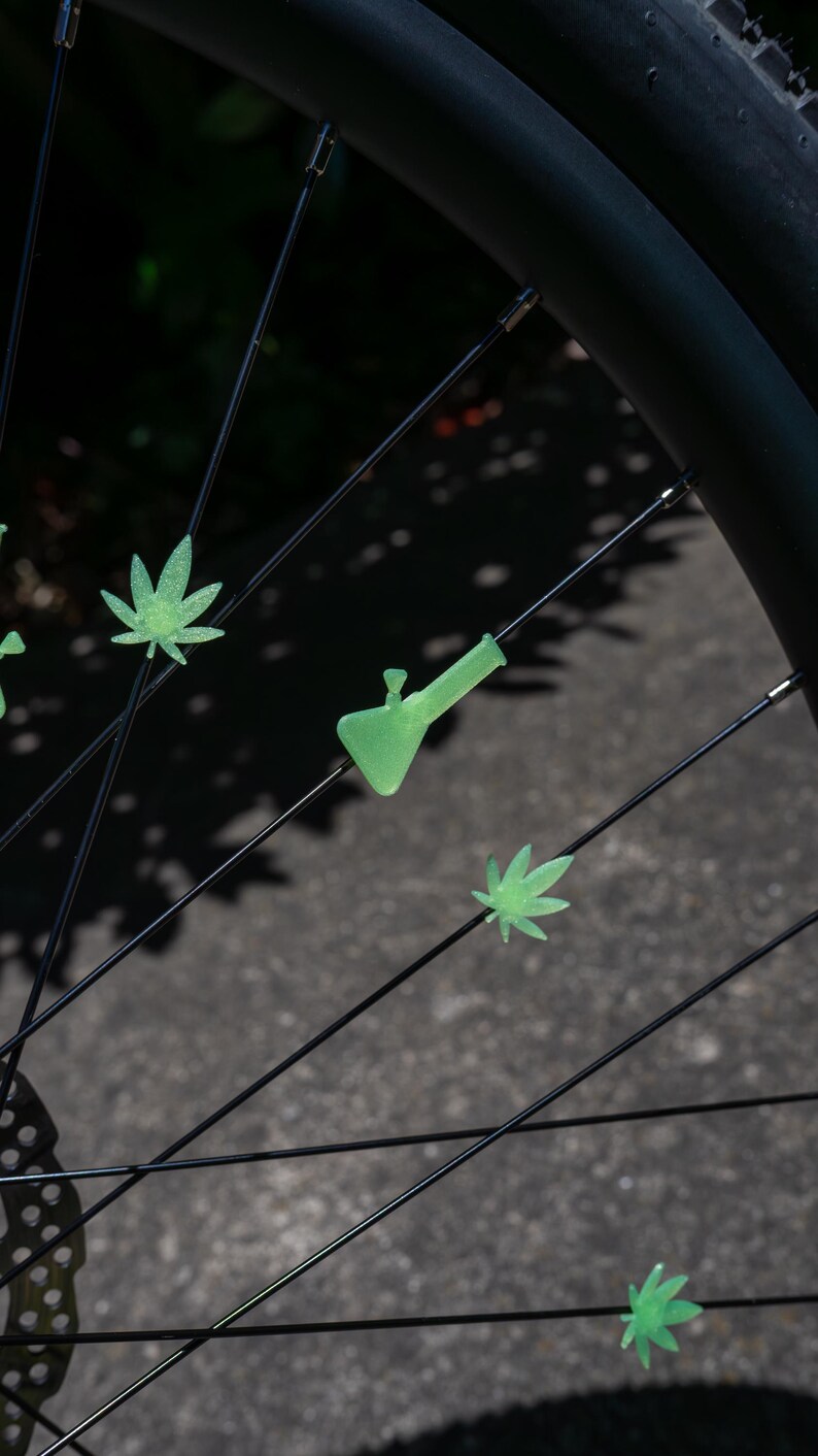 May include: Light green, glow-in-the-dark plastic spoke beads shaped like cannabis leaves and a water pipe attached to the thin black metal spokes of a bicycle wheel. The decorative accessories are arranged individually along the wheel spokes.