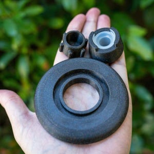 May include: A collection of dark gray, 3D-printed components, including a circular piece with concentric rings, a small cylindrical part with protrusions, and a conical piece with a white interior. The items are held in a hand against a blurred green background.
