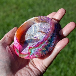 May include: A round, shallow resin dish featuring a vibrant, multi-colored swirl pattern in shades of pink, purple, orange, and blue. The surface has a glossy finish and includes the text ASCENDINGTOM.CO printed in the center of the basin.