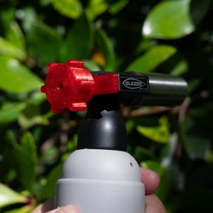 Puede incluir: Un soplete de butano portátil con base gris claro, cuello negro y boquilla metálica. El dispositivo incluye una perilla de ajuste roja brillante, con purpurina y forma de estrella en el lateral con la marca Blazer en una etiqueta negra.
