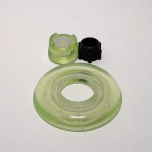 May include: A set of three components including a large, translucent, pale green ring with embedded metallic flecks, a smaller matching pale green cylindrical cap with cutouts, and a small, black, multi-ribbed cylindrical plastic insert.