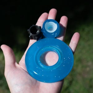 May include: A set of three components including a large translucent blue ring with embedded glitter, a smaller translucent blue circular piece with a white inner core, and a small black cylindrical part with four protruding vertical fins.