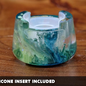 May include: A cylindrical resin holder featuring a swirling blend of teal, blue, and lime green pigments with a translucent finish. The top has a circular cutout and a white silicone insert. The text SILICONE INSERT INCLUDED is at the bottom.