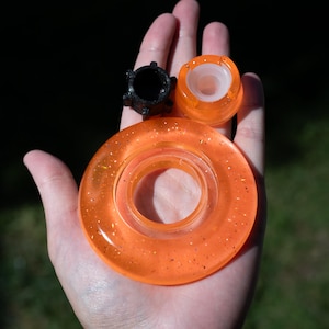 May include: A set of three resin components including a large orange circular ring with embedded glitter, a smaller translucent orange circular piece with a white center, and a small black cylindrical piece with a textured outer edge.