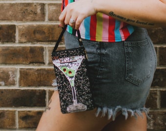 Puede incluir: Un bolso negro de cuentas con forma de copa de martini con aceitunas verdes. El bolso tiene una correa de cadena plateada y lo lleva una persona que lleva una camisa a rayas arcoíris y pantalones cortos de mezclilla.