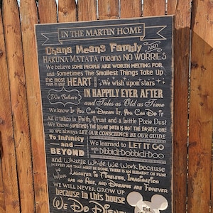 May include: A black wooden sign with gold lettering, featuring a quote about family and Disney. The sign reads "In the Martin Home, Ohana Means Family and Hakuna Matata means No Worries. We believe some people are worth melting for and sometimes the smallest things take up the most heart. We wish upon stars. We believe in happily ever after and tales as old as time. We know if you can dream it, you can do it. All it takes is faith, trust and a little pixie dust. We know sometimes the right path is not the easiest one so we always let our conscience be our guide. To infinity and beyond with a Dibbidi-bobbidi-boo. In every job that must be done, there is an element of fun! And whistle while we work because we believe laughter is timeless, imagination has no age, and dreams are forever. We will never grow up because in this house we do Disney. Est. 10.23.22."