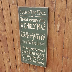Könnte beinhalten: Ein grünes Holzschild mit weißem Text, der lautet: "Code of the Elves Treat every day like Christmas There's room for everyone on the nice list The best way to spread Christmas cheer is singing loud for all to hear!"