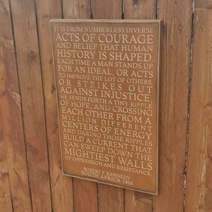 Puede incluir: Un letrero de madera con fondo marrón y texto blanco. El texto dice: "Es de innumerables y diversos actos de coraje y creencia que la historia humana está formada. Cada vez que un hombre se levanta por un ideal, o actúa para mejorar la suerte de los demás, o se opone a la injusticia. Él envía una pequeña ola de esperanza, y cruzándose entre sí desde un millón de centros de energía diferentes y atreviéndose a esas ondas construyen una corriente que puede barrer las paredes más poderosas de la opresión y la resistencia. Robert F. Kennedy Sudáfrica 1966"
