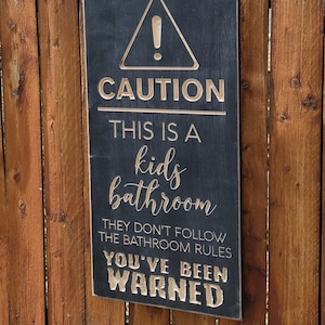 Könnte beinhalten: Rechteckiges Holzschild mit dem Text "CAUTION THIS IS A kids bathroom THEY DON'T FOLLOW THE BATHROOM RULES YOU'VE BEEN WARNED" in einer geschnitzten, hellfarbigen Schrift. Das Schild ist an einem braunen Holzzaun befestigt.