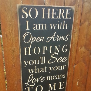 May include: Black wooden sign with white text that reads "SO HERE I am with Open Arms HOPING you'll See what your Love means TO ME Open Arms".