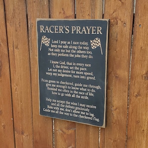 May include: Black wooden sign with gold lettering that reads "Racer's Prayer". The sign features a poem about racing and faith. The sign is mounted on a wooden fence.