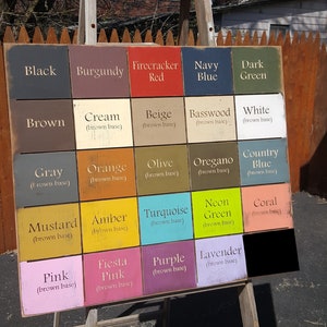 May include: A wooden sign with a grid of 16 painted squares, each with a different color and name. The colors include black, burgundy, firecracker red, navy blue, dark green, brown, cream, beige, basswood, white, gray, orange, olive, oregano, country blue, mustard, amber, turquoise, neon green, coral, pink, fiesta pink, purple, and lavender. Each square has a brown base.