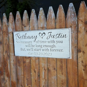 Puede incluir: Un letrero de madera con los nombres "Bethany & Justin" y un símbolo de corazón, montado en una valla de madera. El letrero incluye el texto "No measure of time with you will be long enough. But, we'll start with forever. Est. 03.23.2023."
