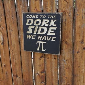 May include: A black wooden sign with white lettering saying "COME TO THE DORK SIDE WE HAVE π" is attached to a wooden fence. The fence is made of vertical wooden planks, with a natural wood finish.
