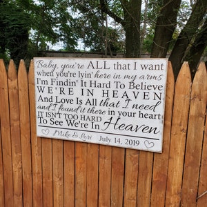 May include: White wooden sign with black text that reads "Baby, you're ALL that I want when you're lyin' here in my arms I'm Findin' It Hard To Believe WE'RE IN HEAVEN And Love Is All that I need and I found it there in your heart IT ISN'T TOO HARD To See We're In Heaven" with a heart symbol before and after the names "Mike & Lori" and the date "July 14, 2019".