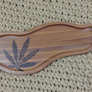 May include: A smooth, brown wooden tray with an irregular, elongated shape and a raised rim. The surface features a dark, wood-burned graphic of a seven-pointed cannabis leaf positioned on the left side, showcasing natural wood grain patterns.