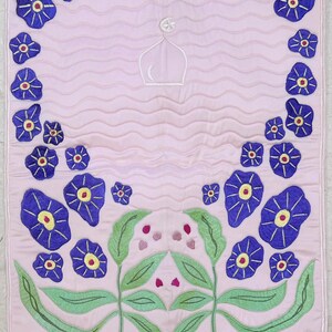 May include: A pink prayer rug with a white mosque design and blue embroidered flowers. The rug has a white lace border.