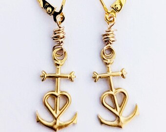 May include: A pair of gold dangle earrings featuring a leverback closure and a charm shaped like an anchor with a heart-shaped centre. The anchor charm is connected to the earring wire by a small gold jump ring and a coiled gold wire detail.