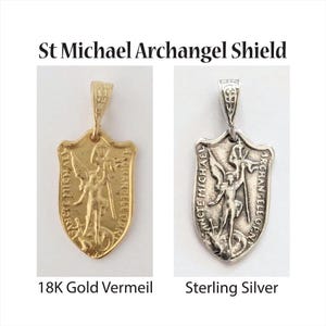 Pode incluir: Dois pingentes em forma de escudo, um dourado e outro prateado, cada um com a imagem de São Miguel. O pingente dourado é rotulado como "18K Gold Vermeil", e o prateado como "Sterling Silver". O texto "St Michael Archangel Shield" está no topo.