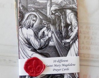 May include: A stack of 10 black and white prayer cards featuring an image of Saint Mary Magdalene. The cards are tied together with a white ribbon and a red wax seal with a fleur-de-lis design. The text on the cards reads "10 different Saint Mary Magdalene Prayer Cards".