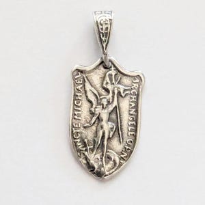 May include: A silver pendant featuring a shield shape with an embossed image of Saint Michael. The pendant has the words "SANCTE MICHAEL ARCHANGELE ORA PRO NOBIS" around the edges. The pendant has a decorative bail.