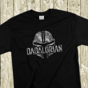 Puede incluir: Camiseta negra con un gr&aacute;fico plateado desgastado de un casco y la palabra "DADALORIAN". El dise&ntilde;o est&aacute; centrado en la parte delantera de la camiseta. La camiseta est&aacute; hecha de un material suave.