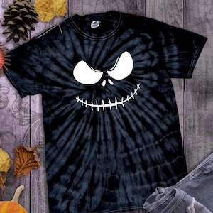 May include: Black tie-dye t-shirt featuring a white Jack Skellington face design. The shirt is paired with blue denim jeans. The image is set against a rustic wooden background with fall-themed decorations.