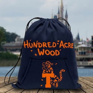 May include: A navy blue drawstring bag with a large orange graphic of Tigger from Winnie the Pooh sitting on a large orange letter T. The bag has a pocket on the front and the text "Hundred Acre Wood" in orange.