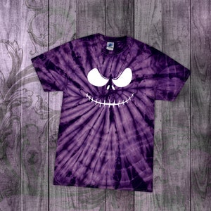 May include: Purple tie-dye t-shirt featuring a white, stylized face design. The face has large, curved eyes and a stitched-mouth grin. The shirt is made of a soft material and has a crew neck. The background is a wooden surface.