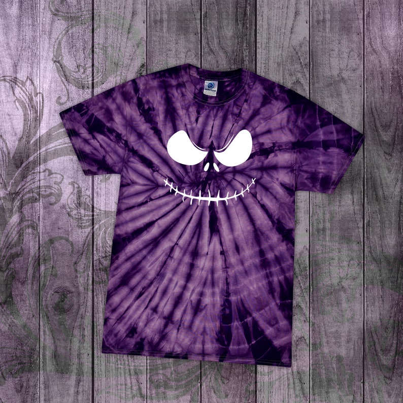 May include: Purple tie-dye t-shirt featuring a white, stylized face design. The face has large, curved eyes and a stitched-mouth grin. The shirt is made of a soft material and has a crew neck. The background is a wooden surface.