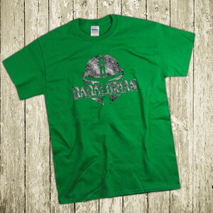 Puede incluir: Camiseta verde con un gr&aacute;fico plateado desgastado de un casco mandaloriano y la palabra "DADALORIAN" impresa debajo. La camiseta es de un material suave y se muestra sobre una superficie de madera r&uacute;stica.