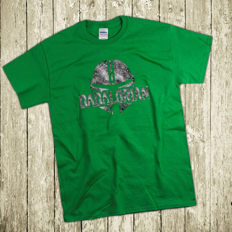 Puede incluir: Camiseta verde con un gr&aacute;fico plateado desgastado de un casco mandaloriano y la palabra "DADALORIAN" impresa debajo. La camiseta es de un material suave y se muestra sobre una superficie de madera r&uacute;stica.