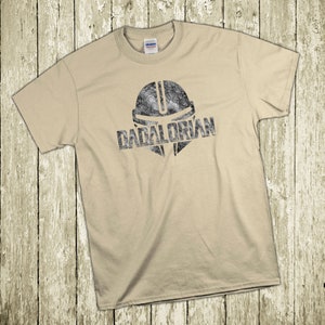 Puede incluir: Camiseta color beige con un dise&ntilde;o negro desgastado de un casco y la palabra "DADALORIAN". La camiseta est&aacute; colocada sobre una superficie de madera clara.
