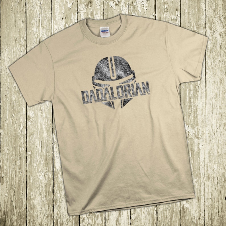 Puede incluir: Camiseta color beige con un dise&ntilde;o negro desgastado de un casco y la palabra "DADALORIAN". La camiseta est&aacute; colocada sobre una superficie de madera clara.