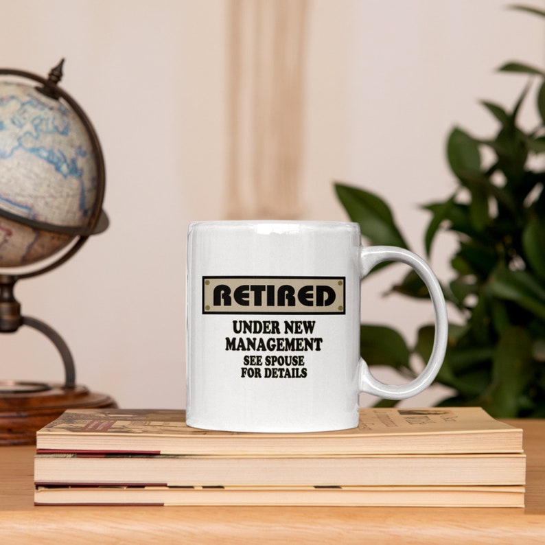 Op de afbeelding: Witte keramische koffiemok met een zwarte en bruine afbeelding met de tekst "Met pensioen Onder nieuw management Zie echtgenoot voor details".
