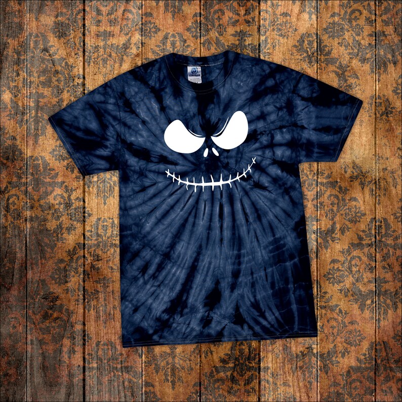 May include: Navy blue tie-dye t-shirt featuring a white, stylized face with large eyes and a stitched grin. The shirt has a classic crew neck and short sleeves. The design is centered on the front of the shirt, creating a spooky, Halloween-themed look.