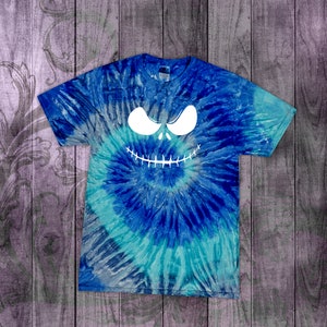May include: A blue tie-dye t-shirt with a white graphic of a spooky face. The face has large, angled eyes and a stitched-mouth grin. The shirt is a swirl of blues and turquoise.