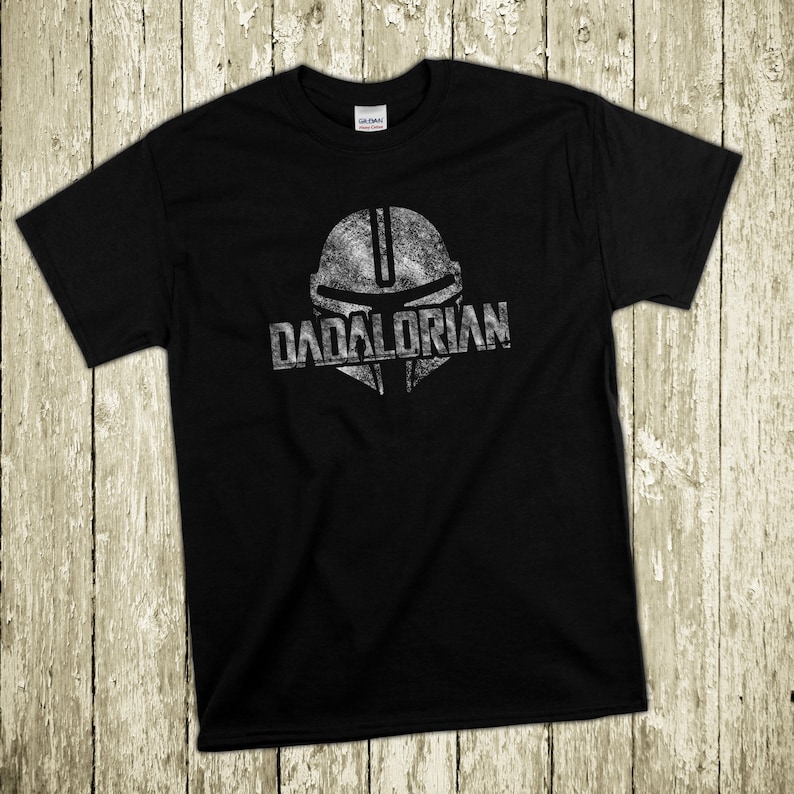 Puede incluir: Camiseta negra con un gr&aacute;fico plateado desgastado de un casco y la palabra "DADALORIAN". El dise&ntilde;o est&aacute; centrado en la parte delantera de la camiseta. La camiseta est&aacute; hecha de un material suave.
