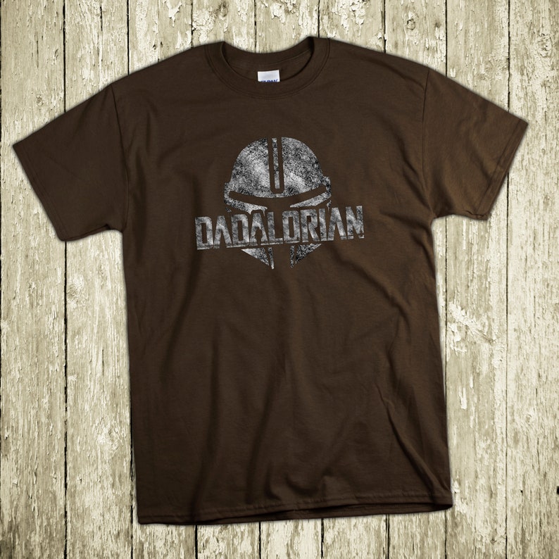 Puede incluir: Camiseta marr&oacute;n con un gr&aacute;fico plateado desgastado de un casco y la palabra "DADALORIAN". La camiseta est&aacute; hecha de un material suave y es adecuada para uso casual. El dise&ntilde;o recuerda a un tema popular de ciencia ficci&oacute;n.