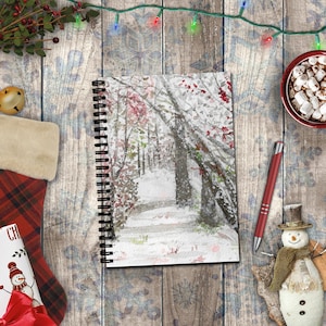 May include: A spiral-bound notebook featuring a watercolor illustration of a snowy forest path with bare trees and hints of red foliage. The cover displays a winter landscape with white snow covering the ground and branches under a soft light.